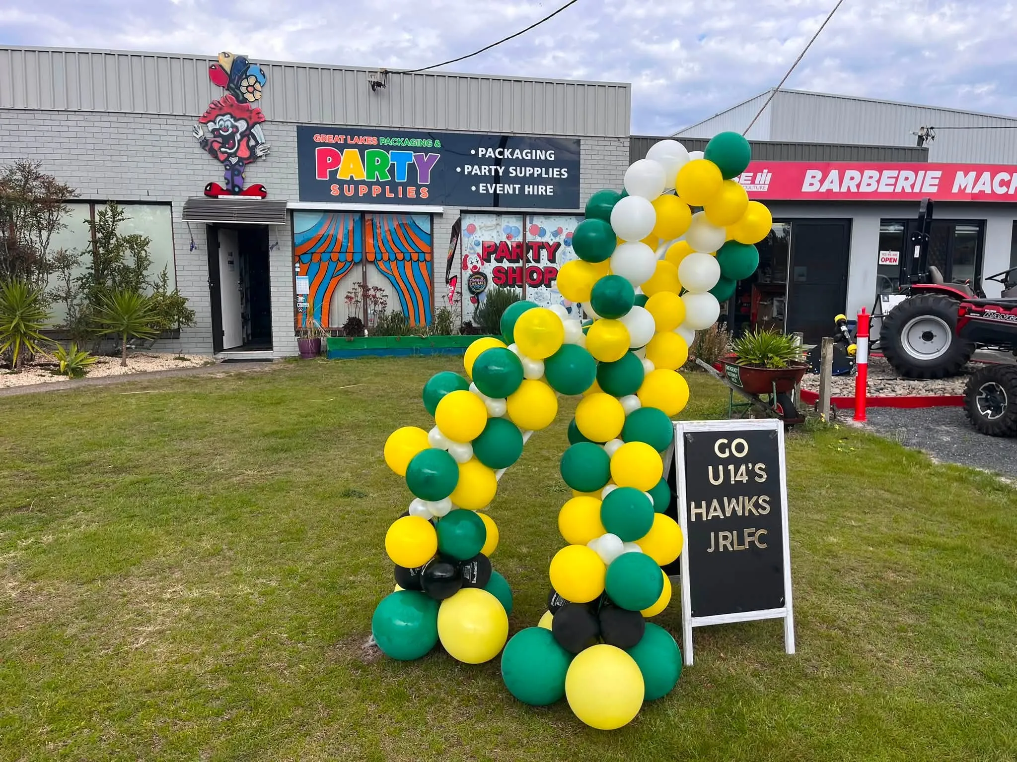 Green, gold and white balloon arch set up outside the store beside a Go U14s Hawks sign